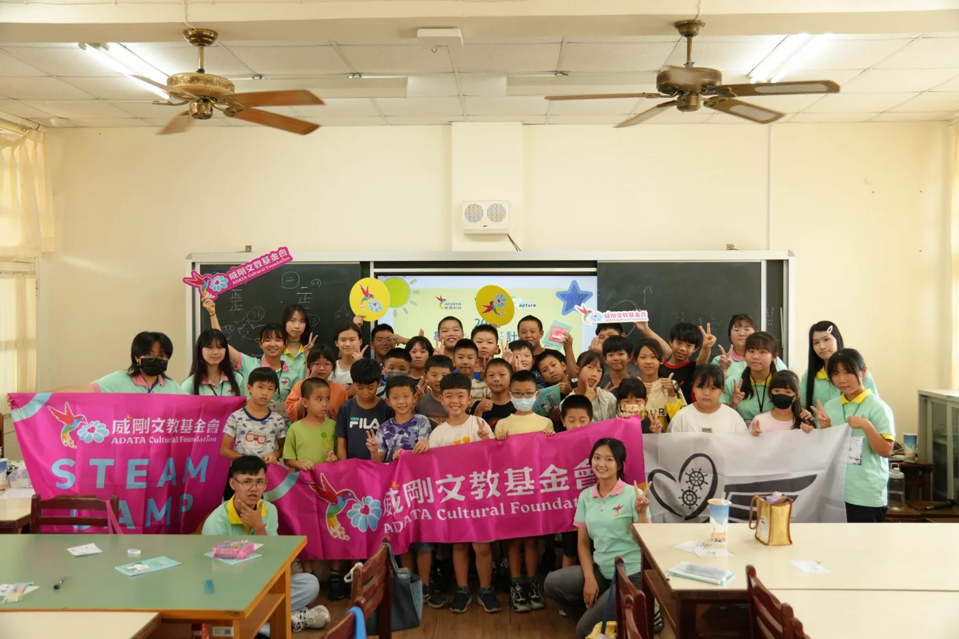 志工與孩子站在教室裡，舉著威剛文教基金會的 STEAM CAMP 旗幟與展心計畫的旗幟合照