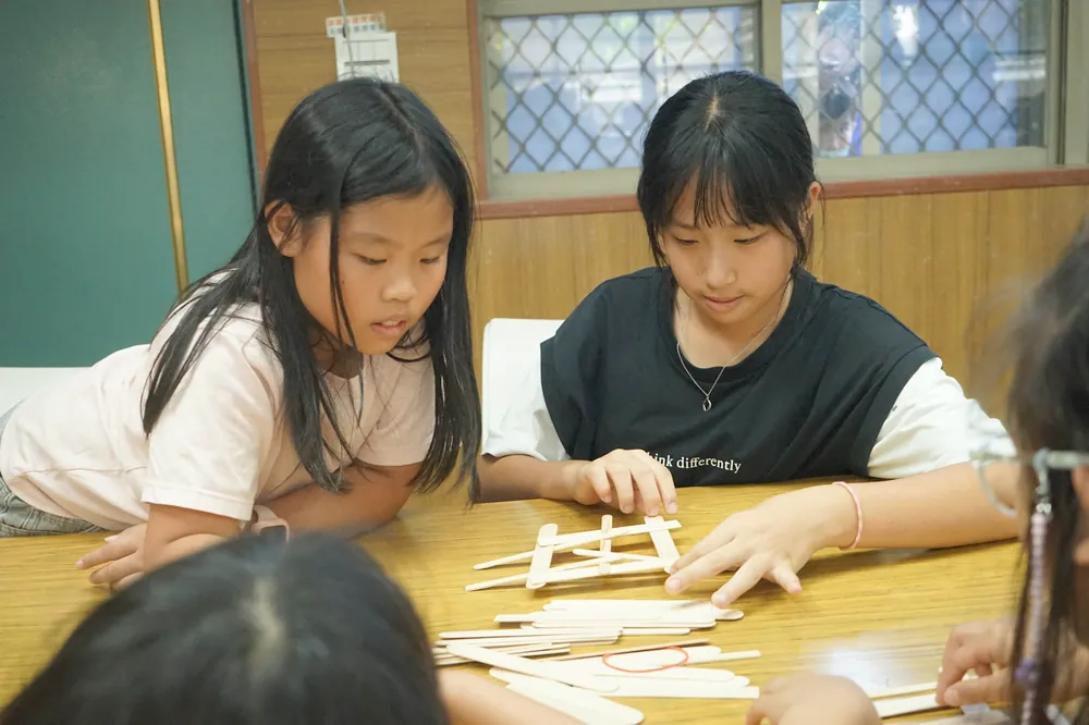 兩個孩子趴在桌邊,專心地用木棍搭橋